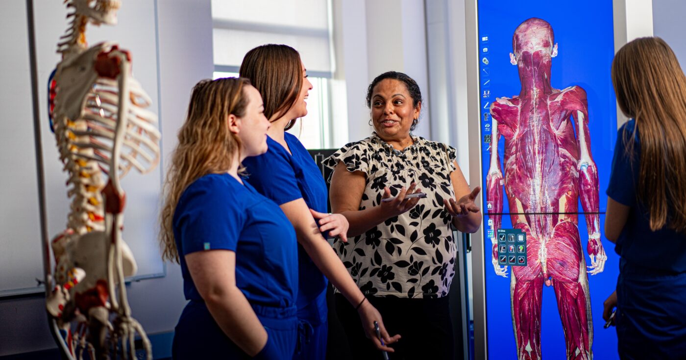 Instructor discussing anatomy with students in blue scrubs beside a digital display of the human muscular system and a skeletal model in a classroom.