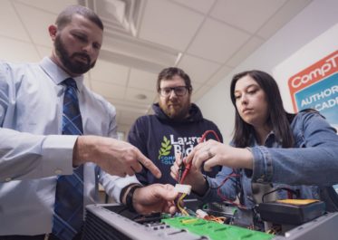 a teacher pointing to a computer circuit board in front of two students
