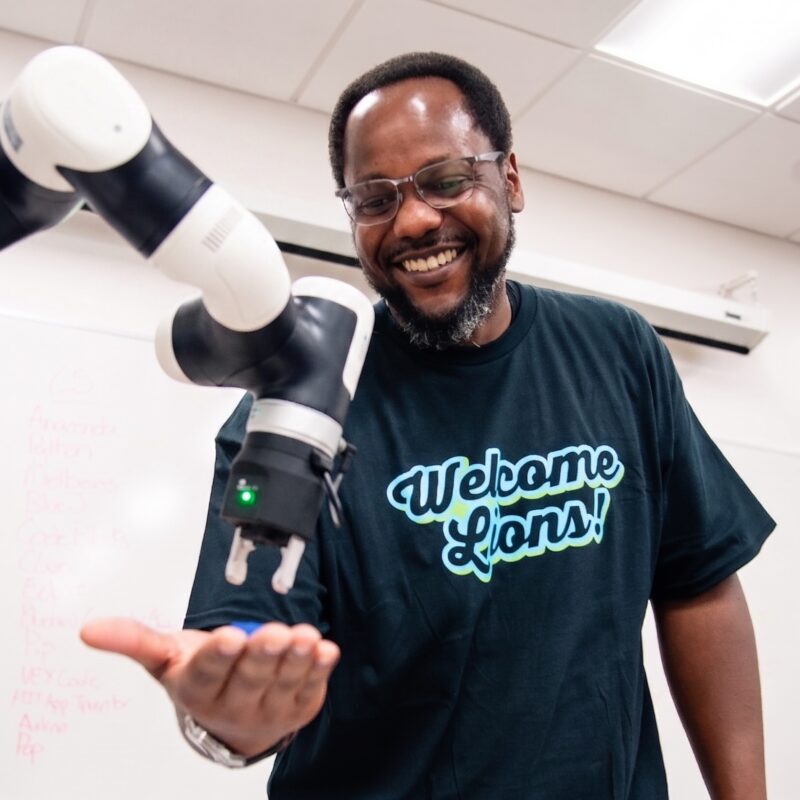 Man in a "Welcome Lions!" T-shirt smiles at a robotic arm reaching toward his open hand.