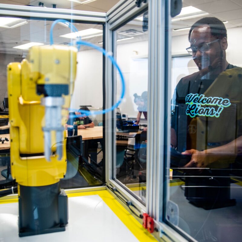 Man in a "Welcome Lions!" T-shirt smiles at a robotic arm reaching toward his open hand.