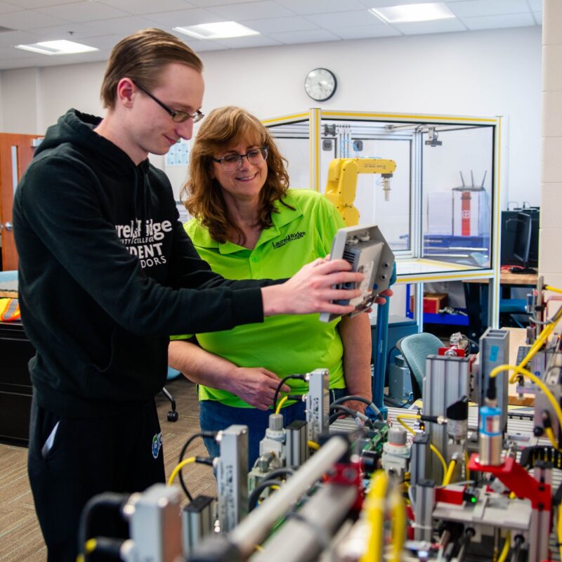 Student and instructor operating a robotic arm control panel in a robotics lab at Laurel Ridge Community College, with a yellow robotic arm and automation equipment in the background.