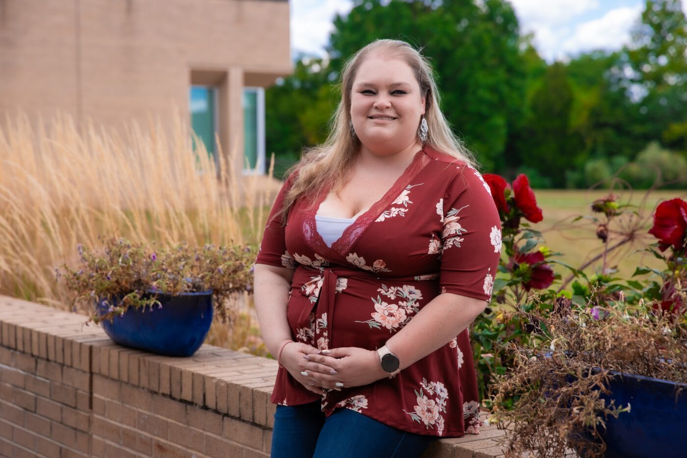 Woman smiling and posing outdoors by a brick wall with plants and flowers, with a building and trees in the background.