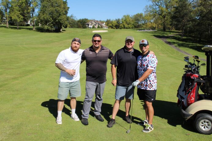 Four men smiling and posing together on a sunny golf course next to a golf cart with clubs.