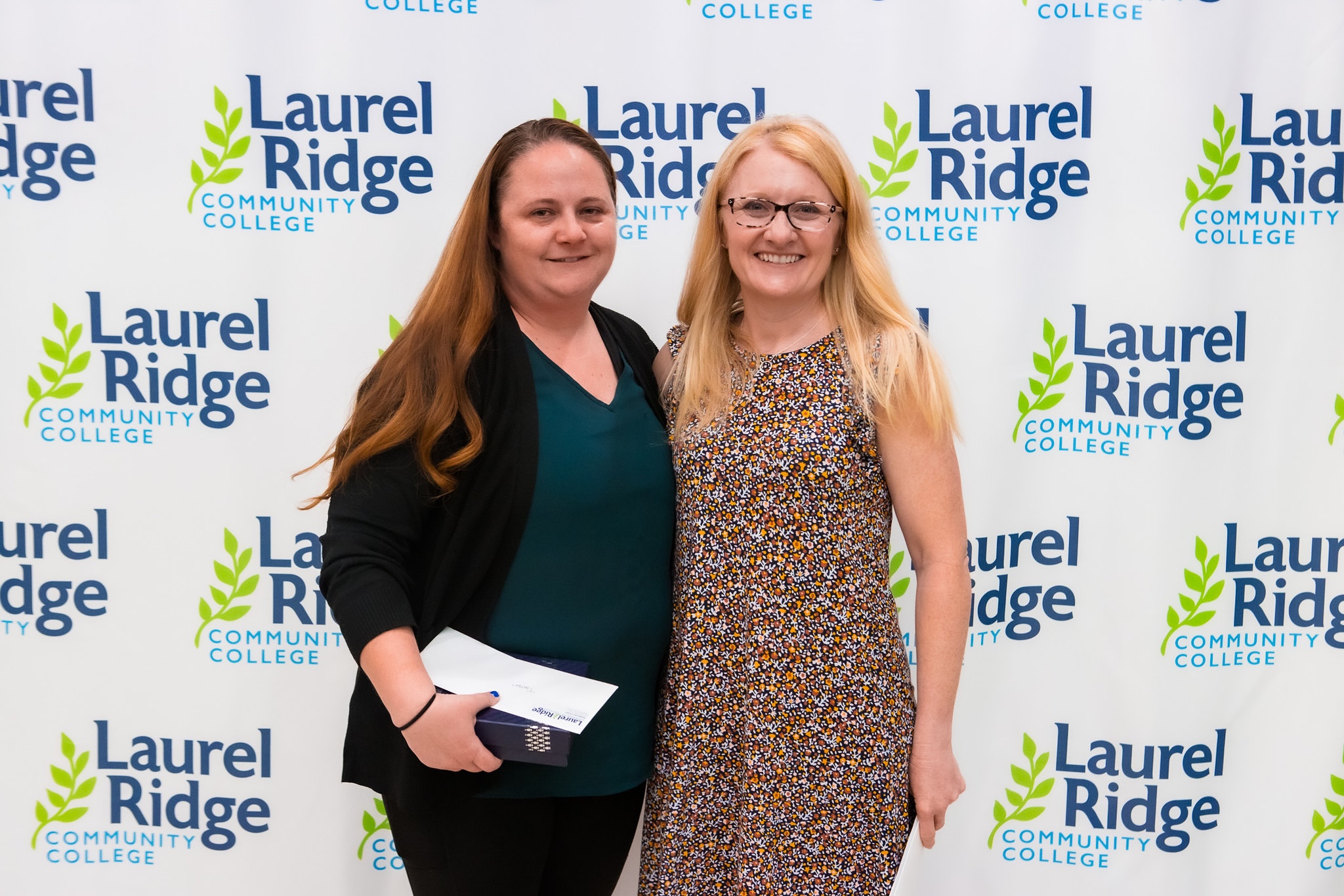 Two women smiling and posing together in front of a Laurel Ridge Community College backdrop, with one holding an envelope and small box.