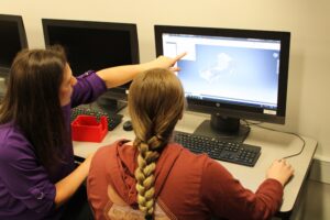 An instructor pointing at a computer screen while helping a student design a 3D model in CAD software.
