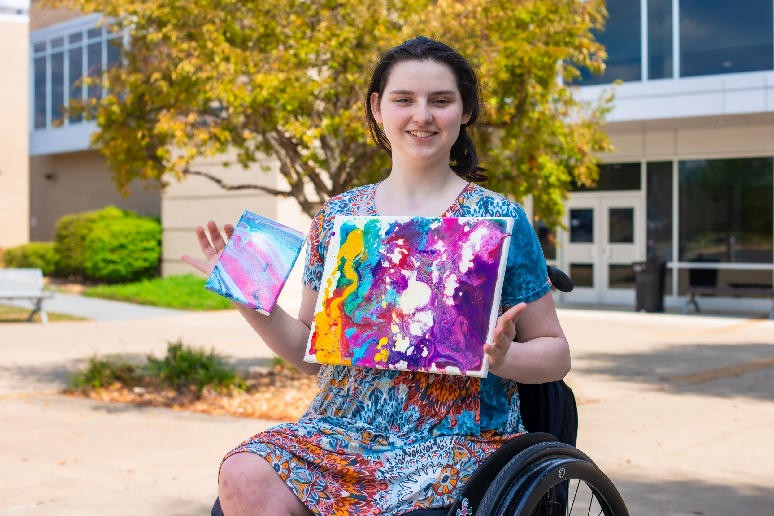 Woman in a colorful dress sitting in a wheelchair outside a building, smiling and holding two abstract acrylic paintings in vibrant colors.