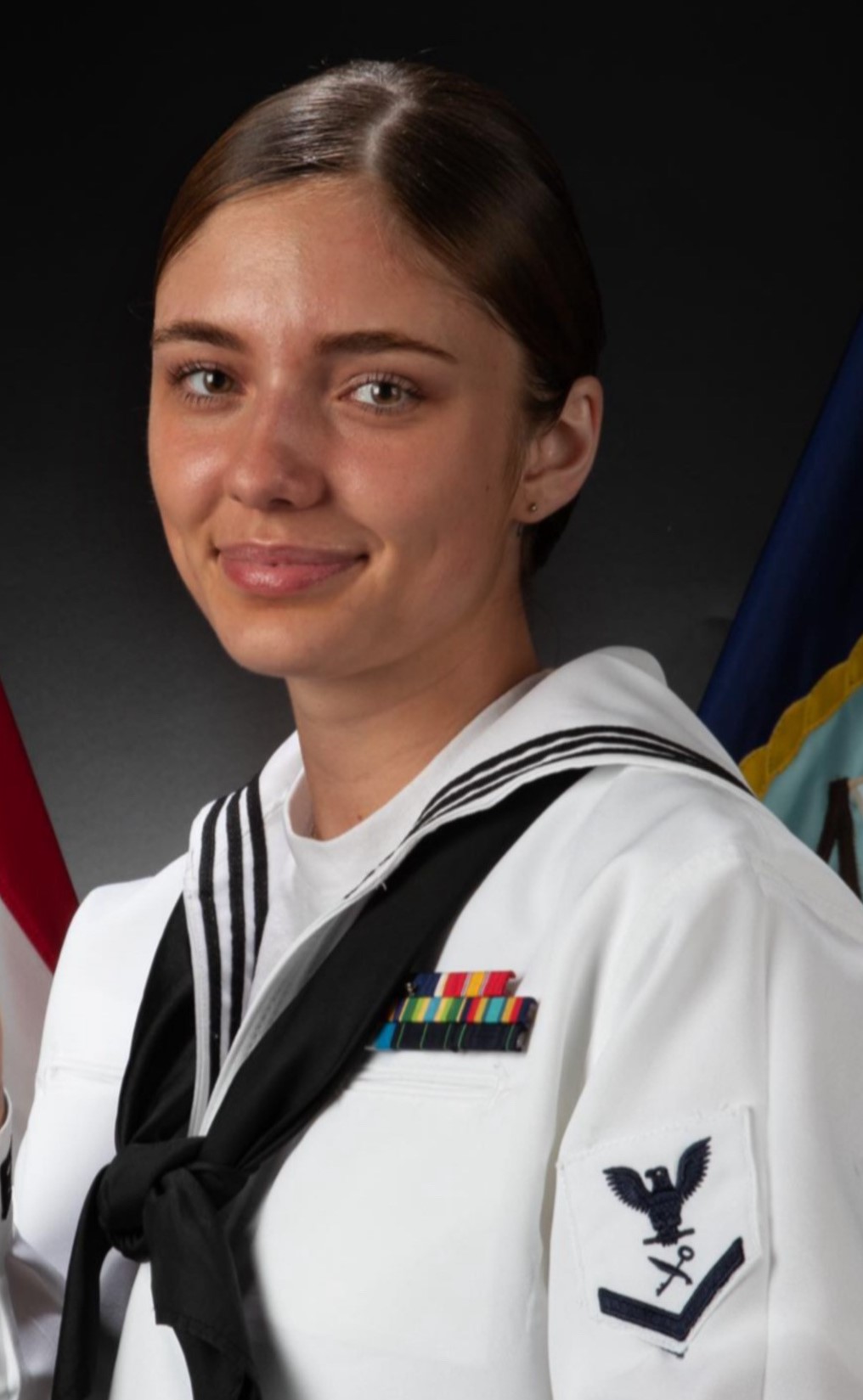 Woman in a U.S. Navy dress white uniform with service ribbons, standing in front of American and Navy flags, smiling slightly at the camera.