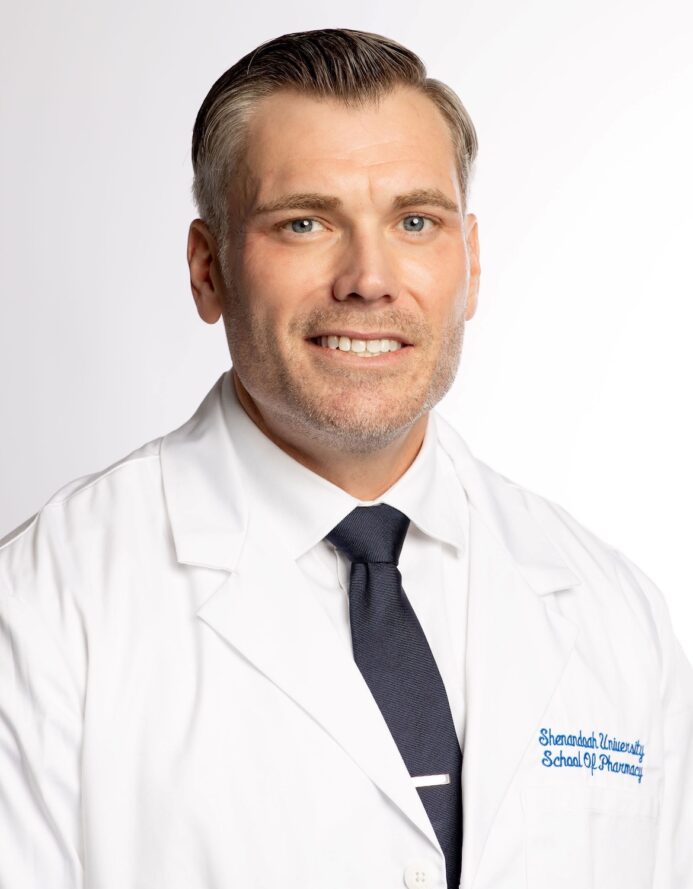 Man wearing a white lab coat embroidered with “Shenandoah University School of Pharmacy,” a white dress shirt, and a dark tie, posing against a plain white background.