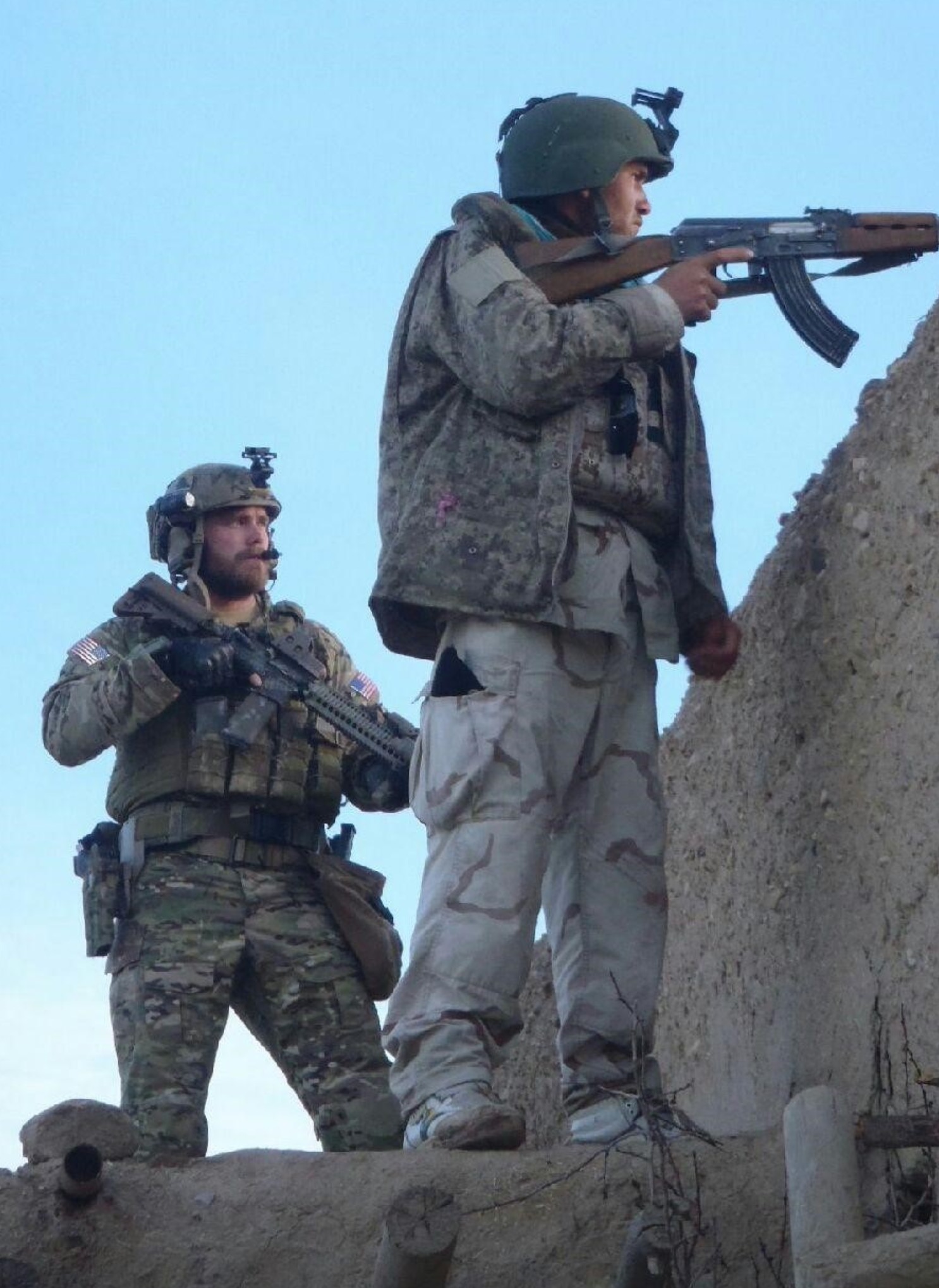 Two soldiers in military gear stand on a dirt structure, one aiming a rifle while the other observes. Both wear helmets with mounted equipment and tactical uniforms, with an American flag patch visible on the gear of the soldier in the foreground.