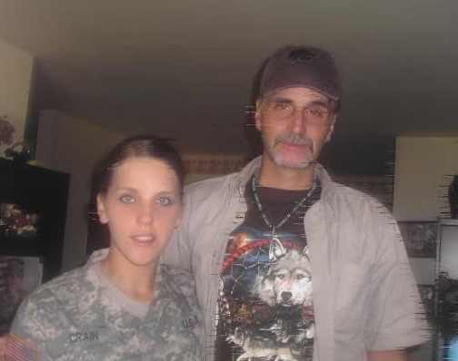 A woman in a U.S. Army uniform stands beside a man wearing a cap and a T-shirt with a wolf graphic, both posing indoors and looking toward the camera.