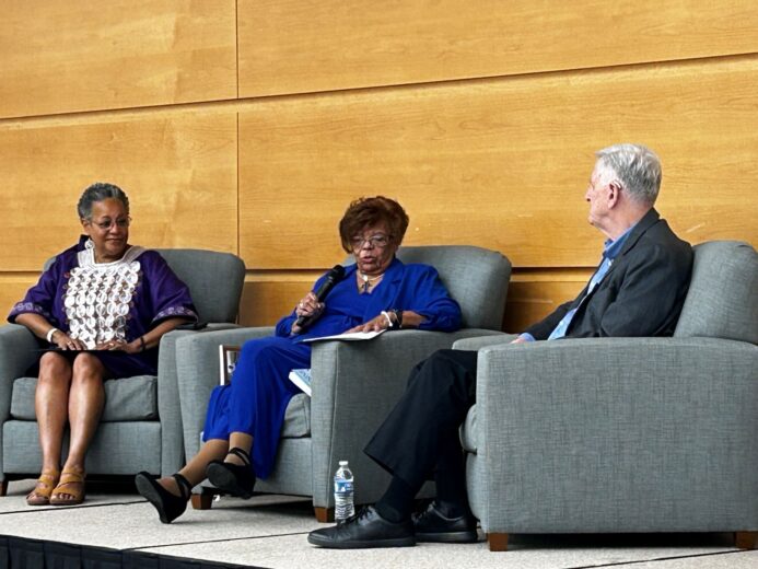Three people seated on stage in armchairs during a panel discussion, with one woman speaking into a microphone while the others listen attentively.
