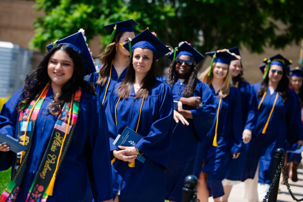 A group of Laurel Ridge Community College graduates in blue caps and gowns walk in a line outdoors, smiling and holding their diplomas during a sunny commencement ceremony.