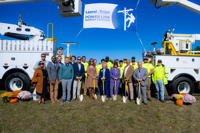 Group of people posing with shovels at a groundbreaking ceremony for the Laurel Ridge Community College Power Line Worker Program, standing between utility trucks under a banner on a clear sunny day.