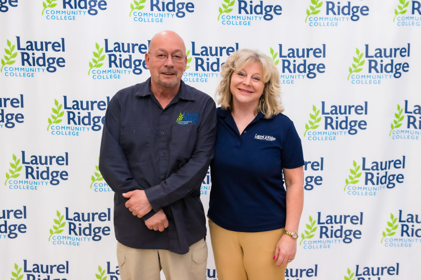 Man and woman smiling and standing together in front of a Laurel Ridge Community College step-and-repeat backdrop, both wearing college-branded shirts.