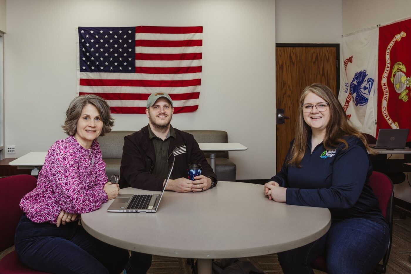Three people sit around a table in a room decorated with American and military flags. One person has a laptop open, suggesting a meeting or study session, and all appear relaxed and engaged.