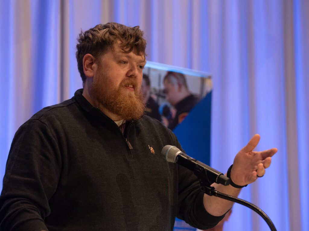 A bearded man is speaking at a podium, gesturing with one hand while standing in front of a microphone. He is wearing a dark long-sleeve shirt and appears to be addressing an audience. Behind him, there is a blurred banner with images of people, and the background features softly lit vertical drapes in shades of blue and purple.