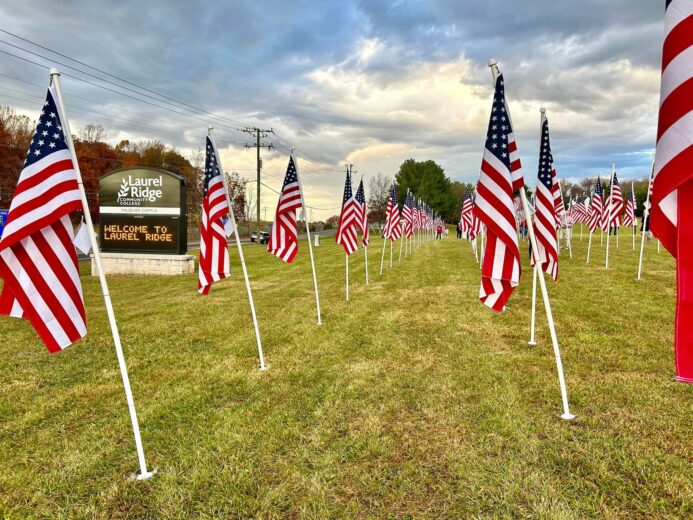 Here is clear, ADA-compliant alt text for the image: **Alt text:** Rows of American flags displayed on poles across a grassy lawn at Laurel Ridge Community College’s Fauquier Campus, with the campus welcome sign visible on the left and a cloudy sky overhead.