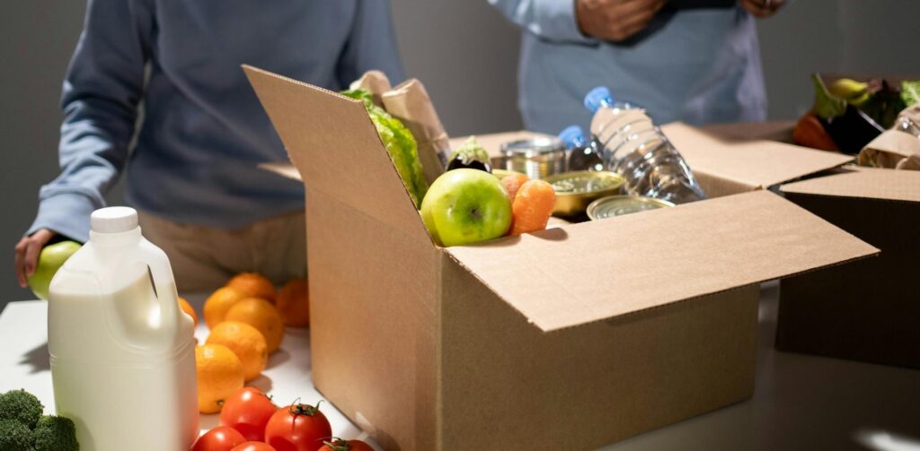 Close-up of volunteers packing food donation boxes with fresh produce, bottled water, canned goods, and other essentials on a table, with a milk jug and vegetables visible in the foreground.