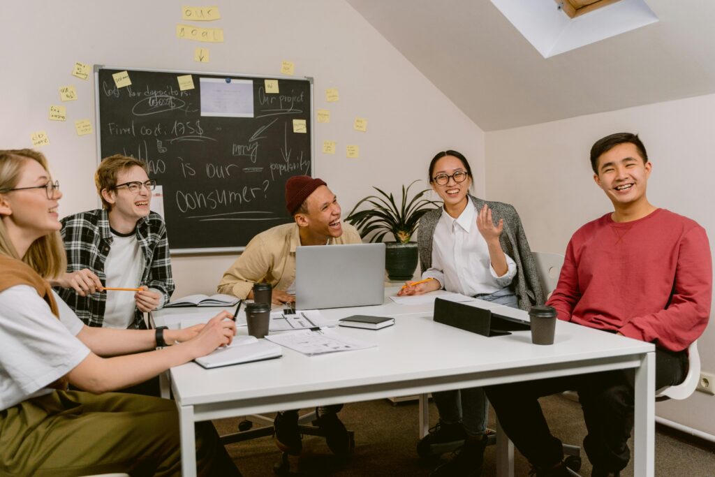 A group of five young adults sit around a white table in a casual meeting room, smiling and laughing together. Notebooks, papers, laptops, and coffee cups are spread across the table. Behind them is a chalkboard covered in notes, diagrams, and sticky notes. The atmosphere appears collaborative and upbeat.