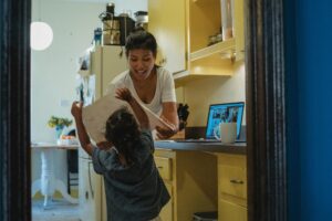 A mother looks at a drawing made by her young daughter while she uses a computer in a kitchen