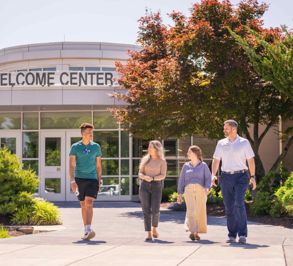 Four people walking together outside a campus welcome center on a sunny day, chatting and smiling in front of trees and landscaped greenery.