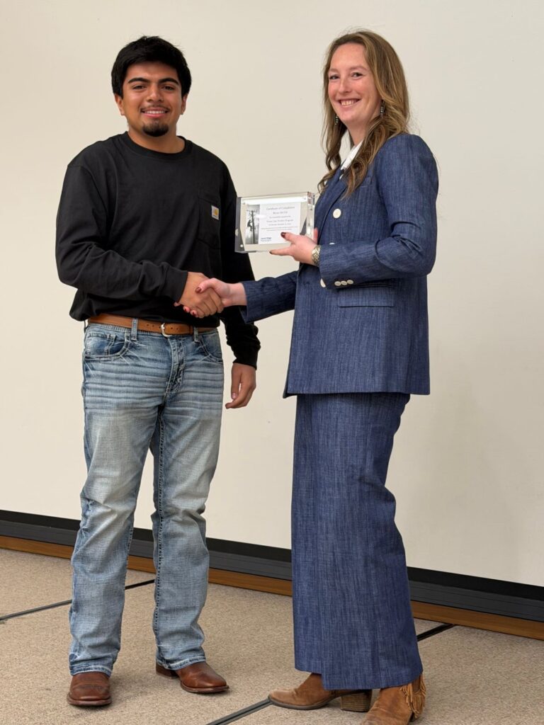 A man stands on a stage shaking hands with a woman holding a plaque. Both are smiling and looking at the viewer.