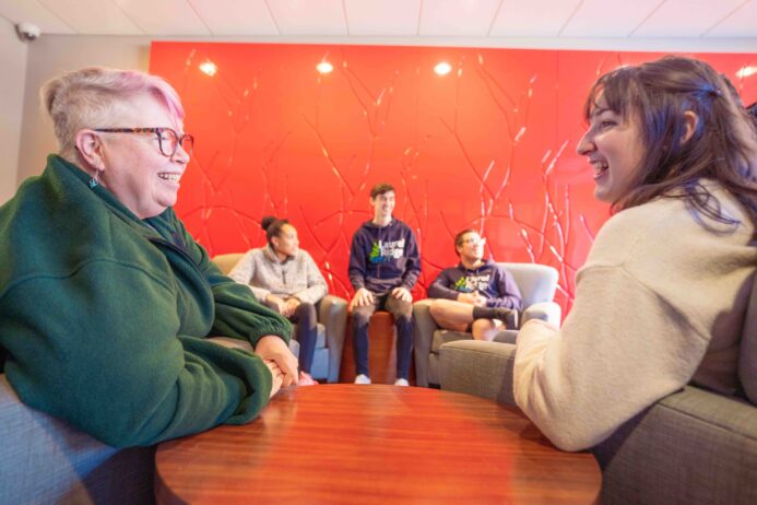 Students sitting and talking in a lounge area with a bright red textured wall, two people conversing in the foreground and three more seated in chairs in the background.