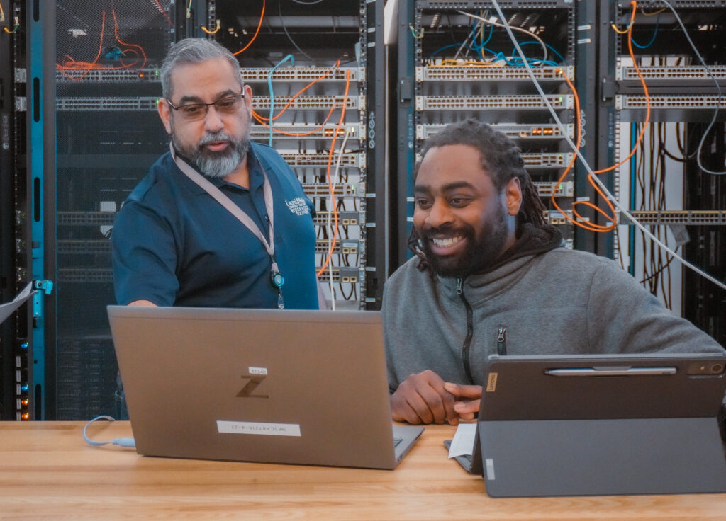 An instructor and student work together at a table reviewing a laptop in a computer networking lab with server racks and cables in the background.