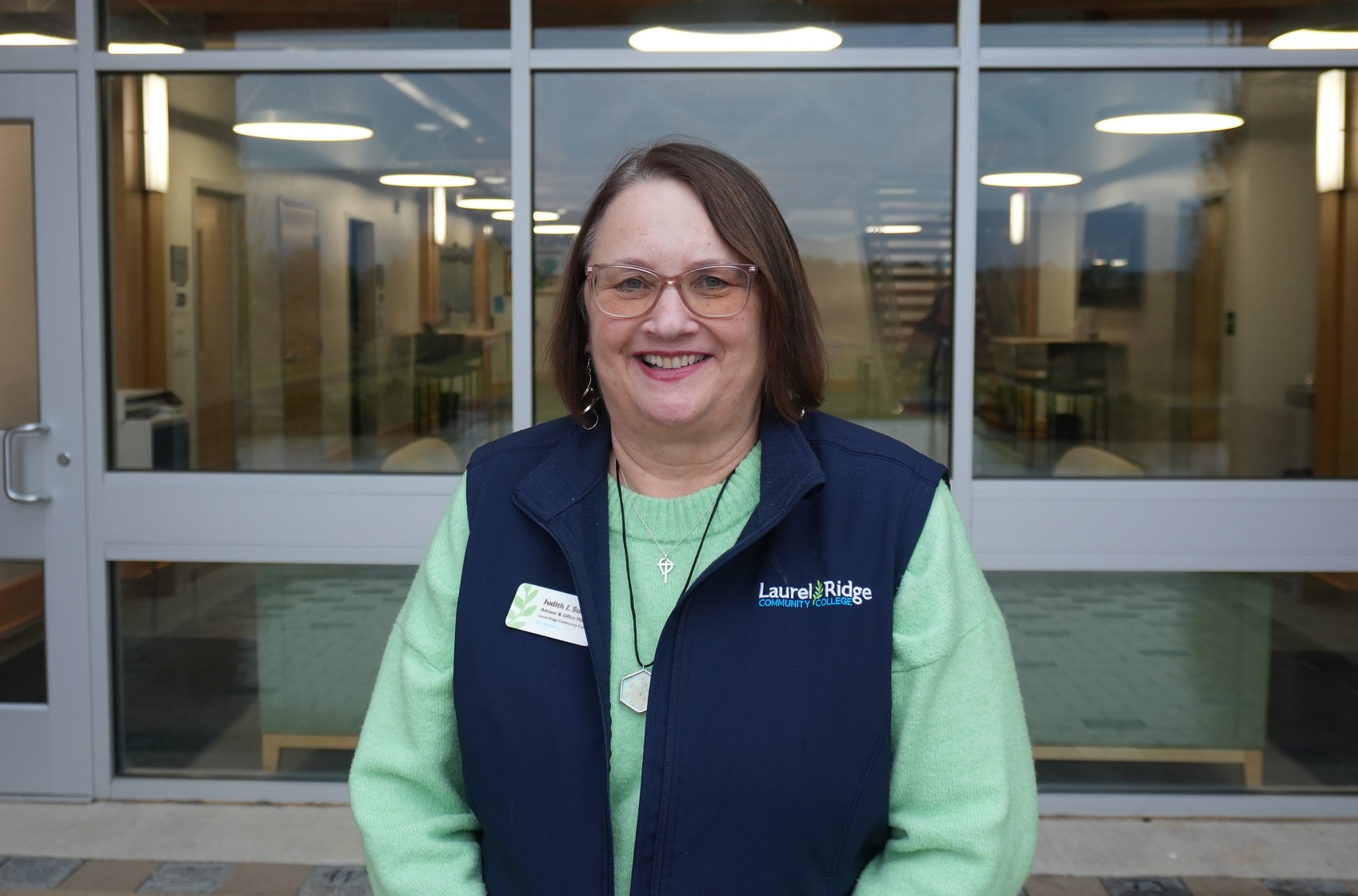Smiling Laurel Ridge Community College staff member Judy Suddith wearing a green sweater and navy vest stands outside the Luray–Page County Center entrance with glass doors behind her.