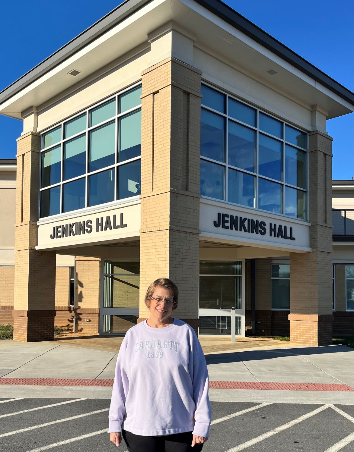 A woman wearing a light-colored sweatshirt stands outside Jenkins Hall, a modern campus building with large windows and brick columns, on a clear sunny day.
