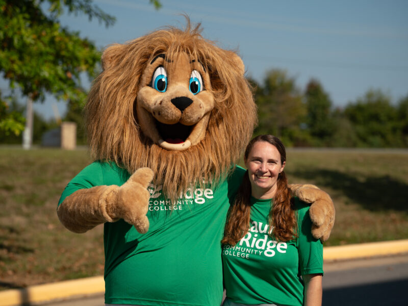Laurel Ridge Community College mascot wearing a green shirt poses with a smiling participant in a matching shirt at an outdoor campus event on a sunny day