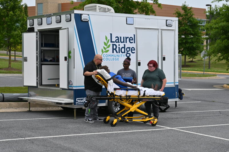 Students practicing an emergency medical scenario outside, loading a patient mannequin on a stretcher beside a Laurel Ridge Community College EMS training ambulance in a parking lot