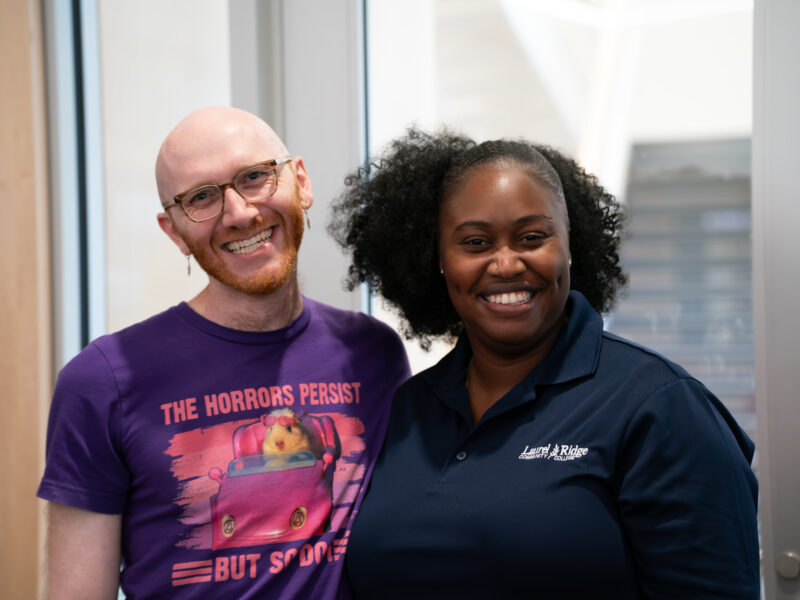 Two smiling Laurel Ridge Community College employees stand side by side indoors, one wearing a purple graphic T-shirt and glasses and the other wearing a navy Laurel Ridge polo shirt, posing together in a bright office hallway