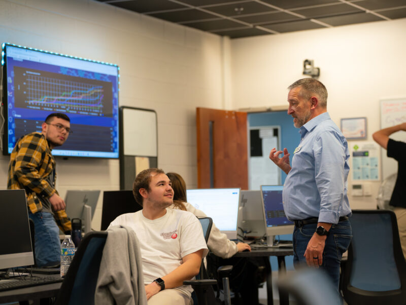 Instructor speaking with students in a computer lab, standing beside workstations while students sit at desktop computers, with a large screen displaying data charts and graphs in the background