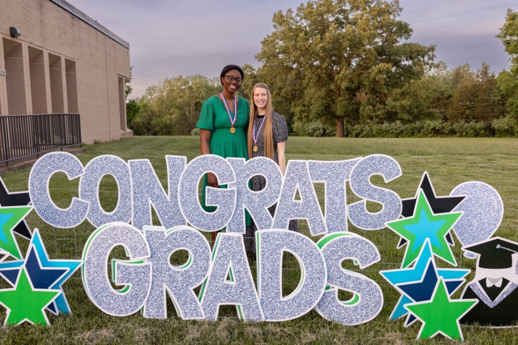 Two graduates wearing medals stand smiling behind a large outdoor “CONGRATS GRADS” sign with star decorations on a grassy campus lawn.