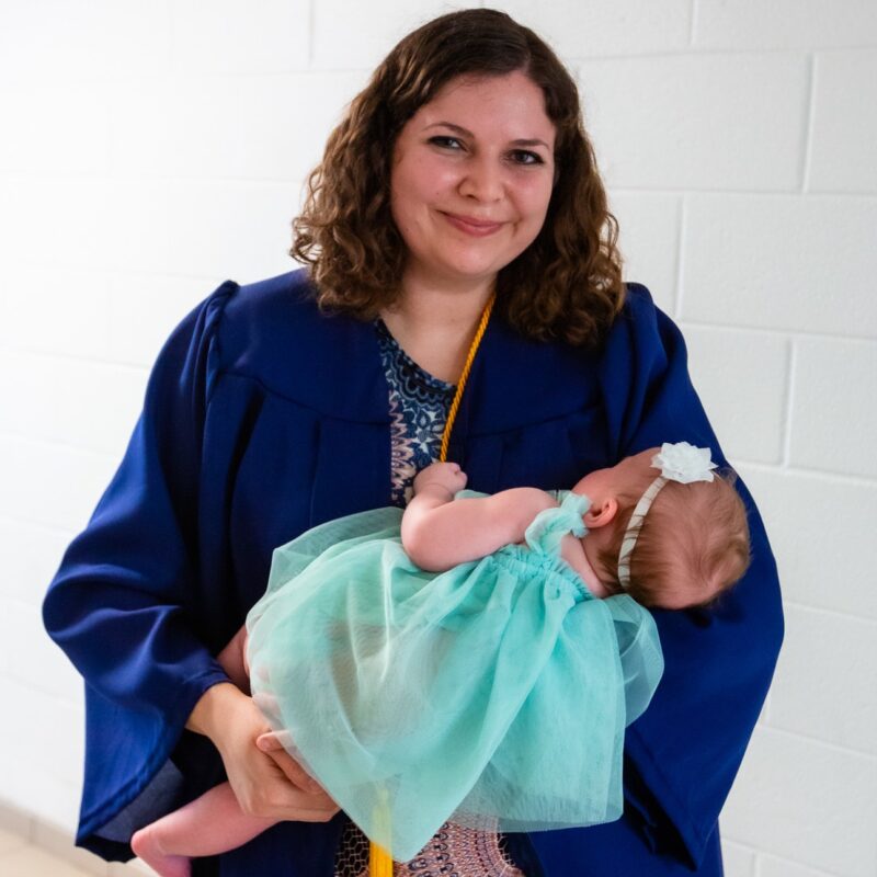 Graduate in blue cap and gown holding a sleeping baby wearing a mint green dress and white headband indoors