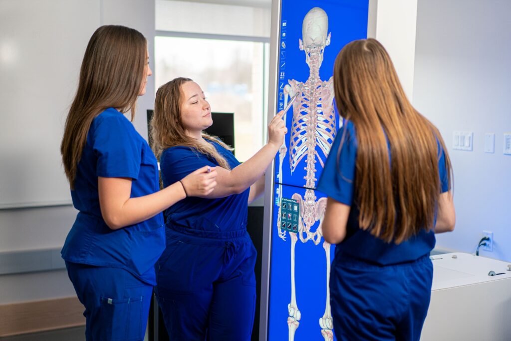 Three physical therapy assistant students in blue scrubs studying a full-body skeletal anatomy image on a large vertical touchscreen display in a classroom.