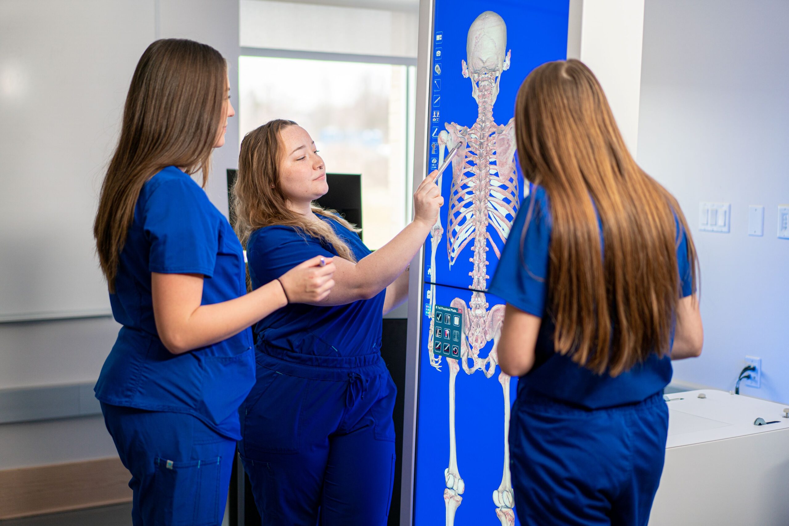 Three physical therapy assistant students in blue scrubs studying a full-body skeletal anatomy image on a large vertical touchscreen display in a classroom.