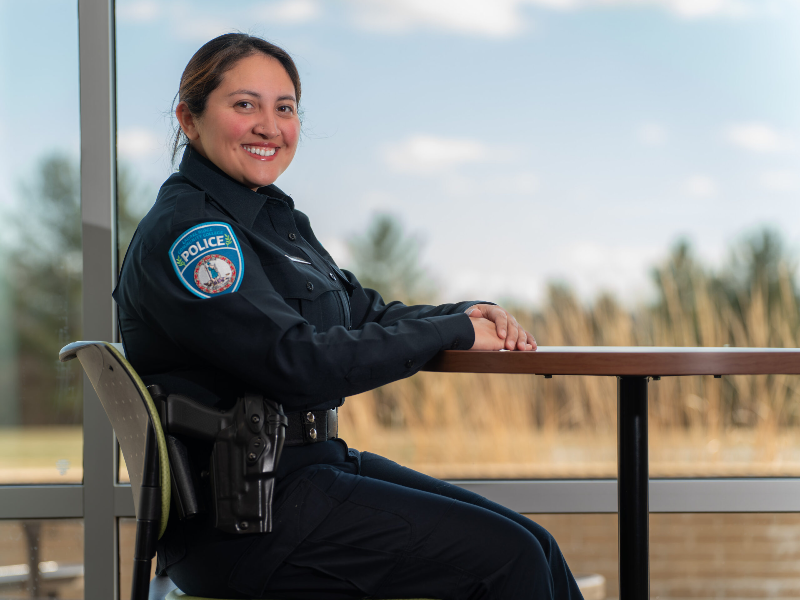 A uniformed police officer sits at a table indoors, smiling toward the camera with a windowed landscape in the background.