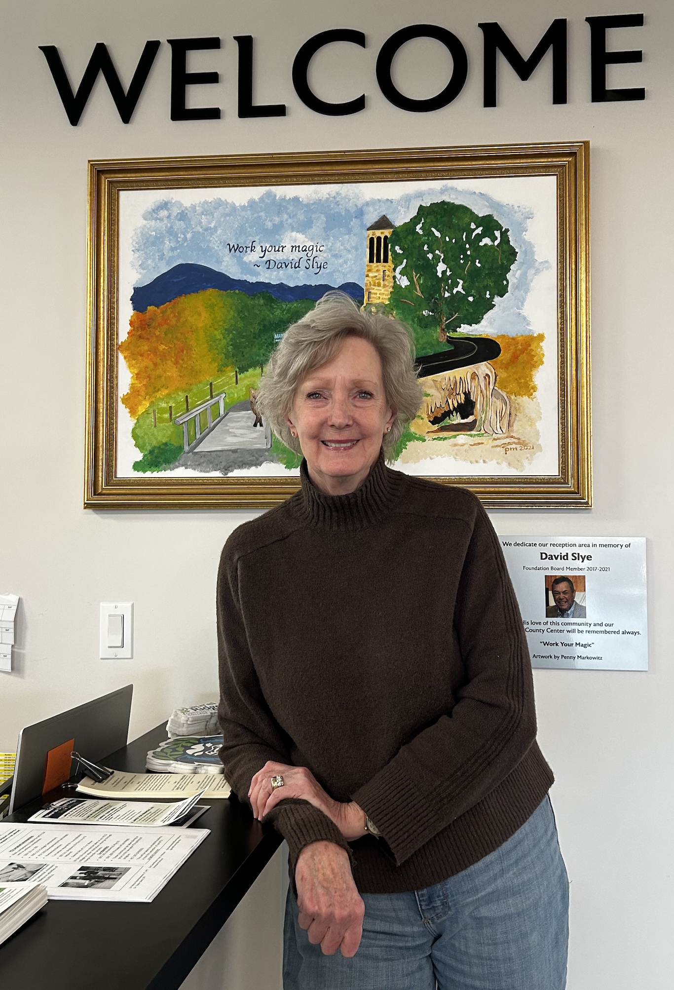 Photograph of Donna leaning against the front desk at Jenkins Hall with a welcome sign behind her