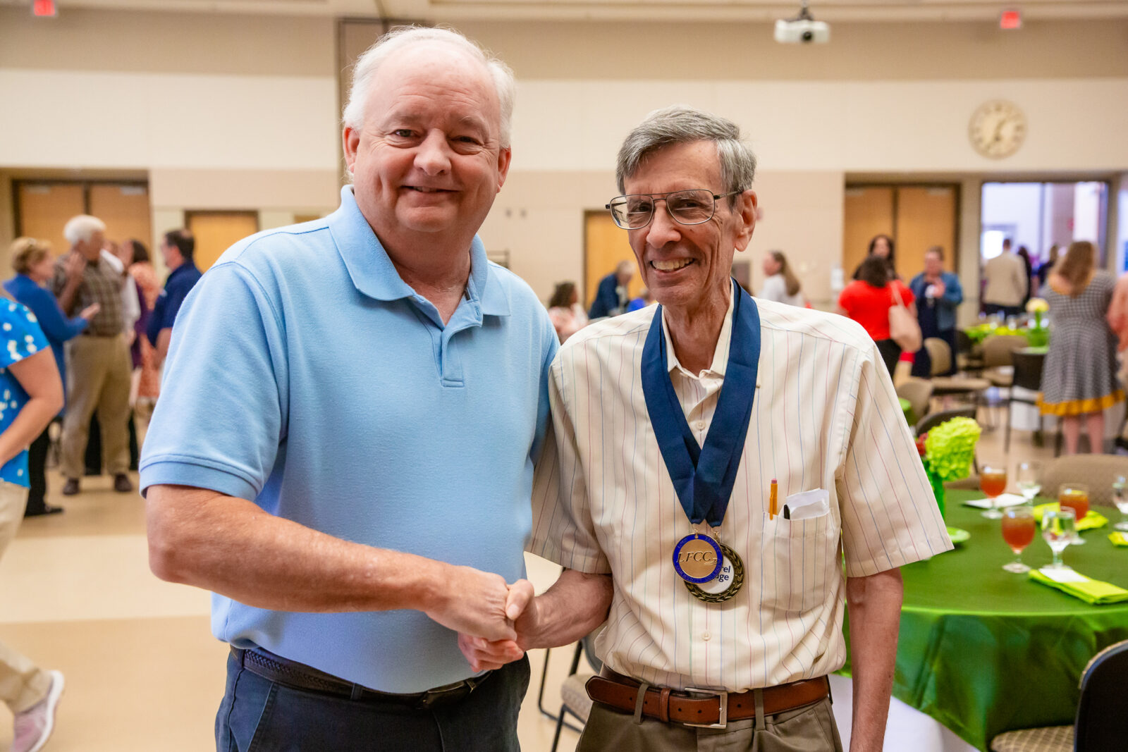 Two men shake hands and smile towards the camera. The man on the right wears two awarded medallions.