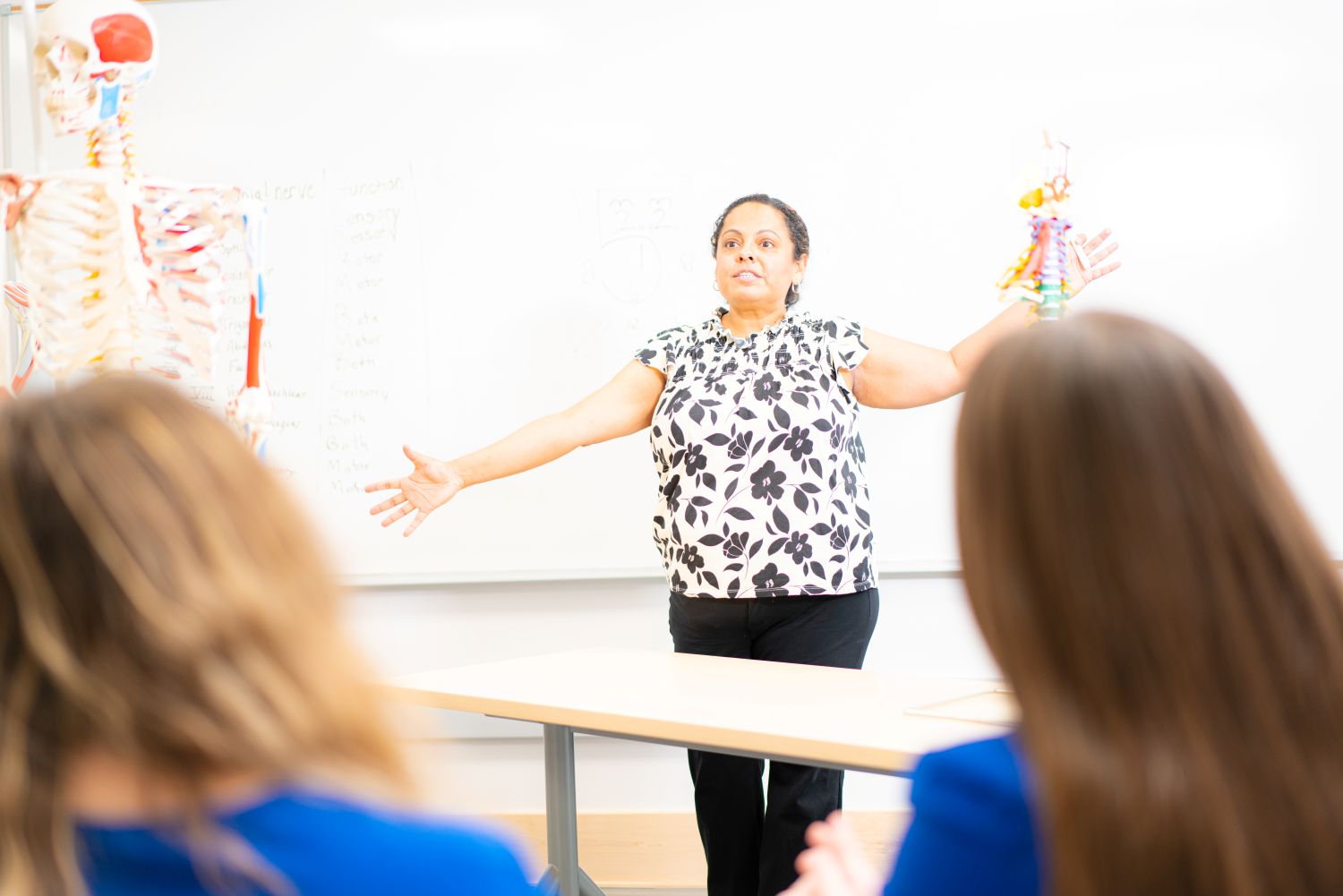 Instructor gestures while teaching anatomy to students in a classroom with a skeleton model visible.