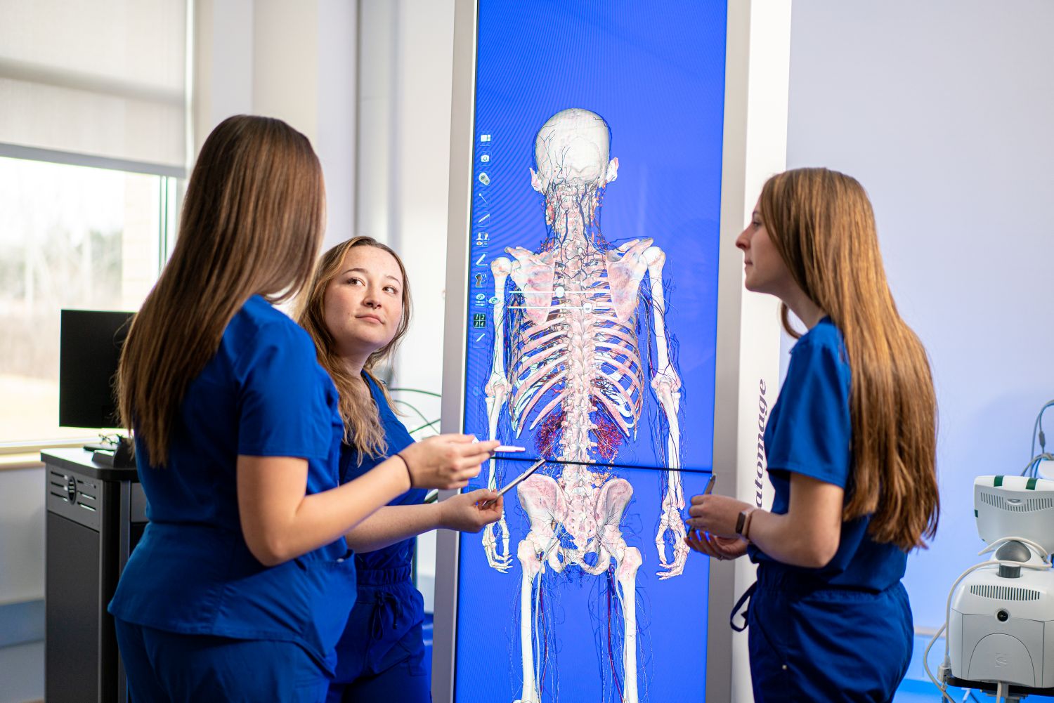 Students in scrubs study a digital human skeleton and anatomy display in a classroom lab.