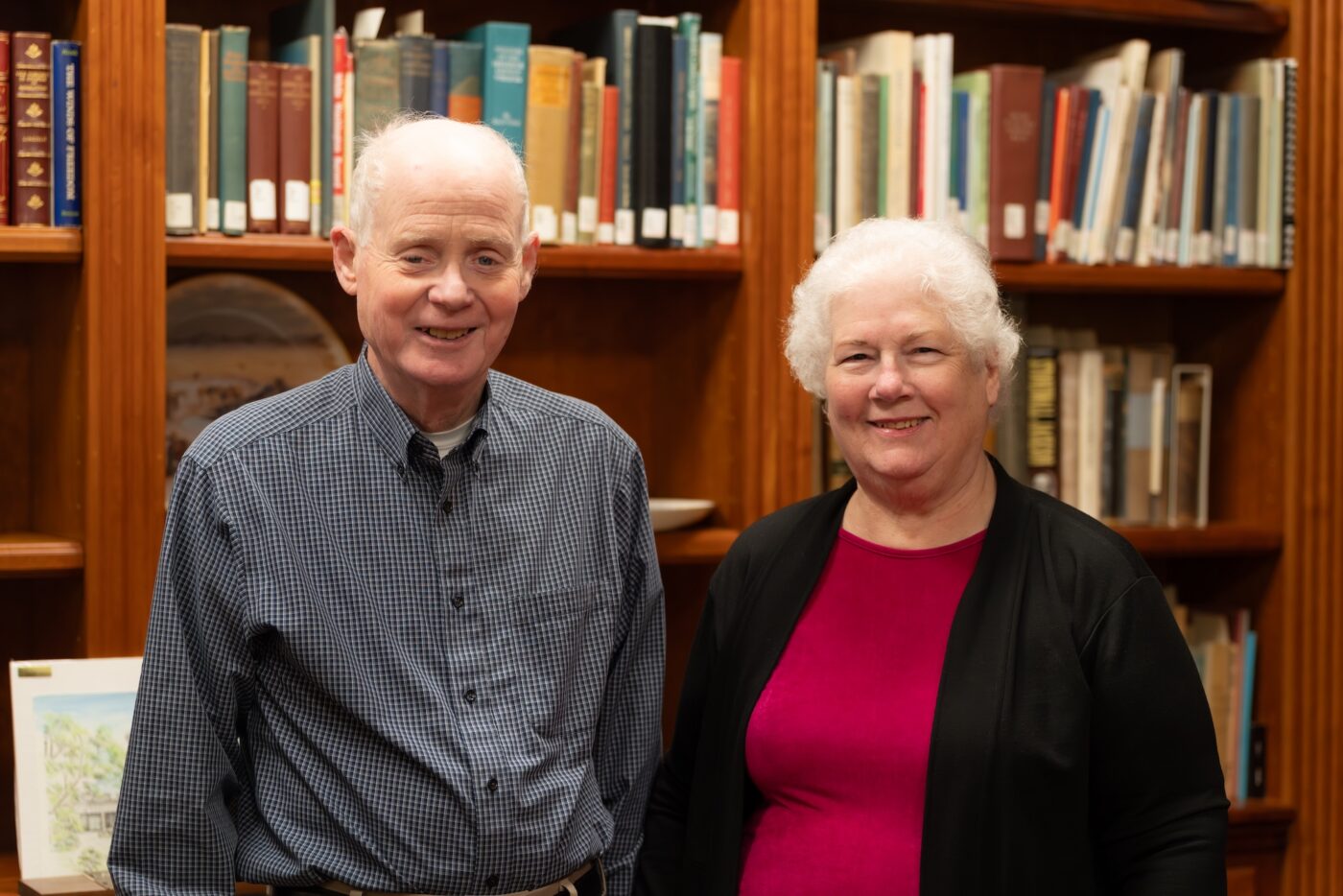 Foss and Margaret Smith smile together in front of a wooden bookshelf filled with books in a library setting. Foss wears a blue plaid button-down shirt and Margaret wears a magenta top with a black cardigan.