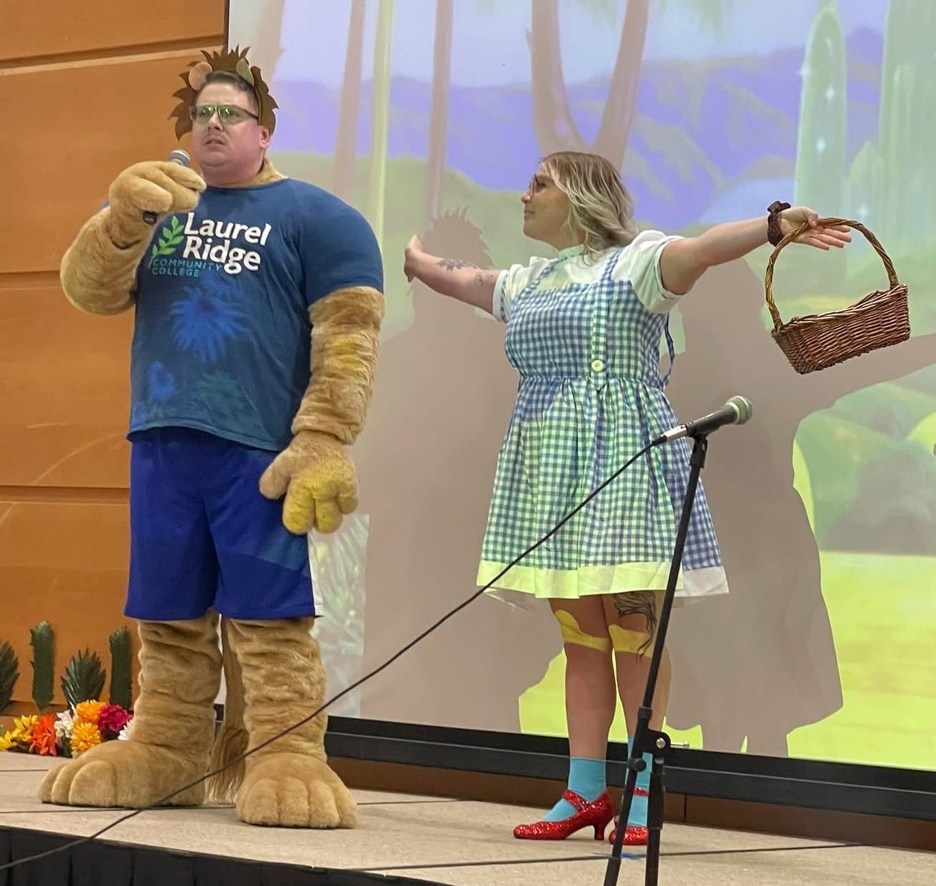 A person dressed as the Cowardly Lion wearing a Laurel Ridge Community College t-shirt, blue shorts, and oversized furry lion paws and feet stands on a stage holding a microphone, next to a person dressed as Dorothy from The Wizard of Oz in a blue gingham dress, red ruby slippers, and blue socks, holding a wicker basket with one arm outstretched, performing in front of a projected Emerald City forest backdrop