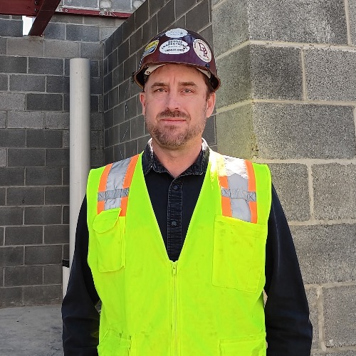 a head shot photo of Jason. He stands amount a concrete structure while wearing a safety helmet and vest.