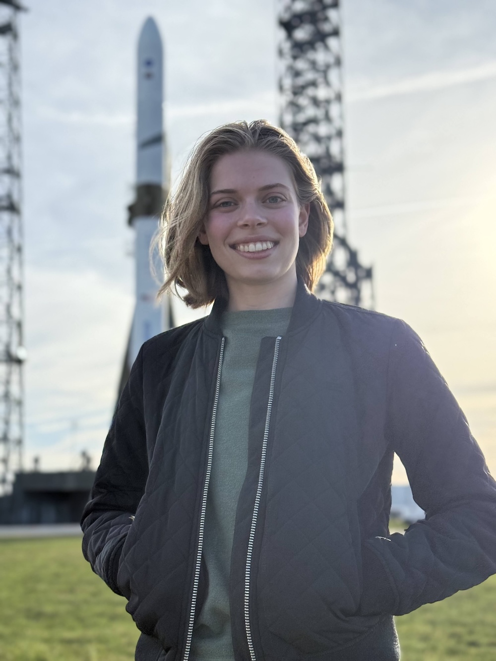 Katie Borland smiles with hands in pockets, wearing a quilted bomber jacket over a green shirt, standing outdoors in front of a rocket on a launch pad at golden hour.