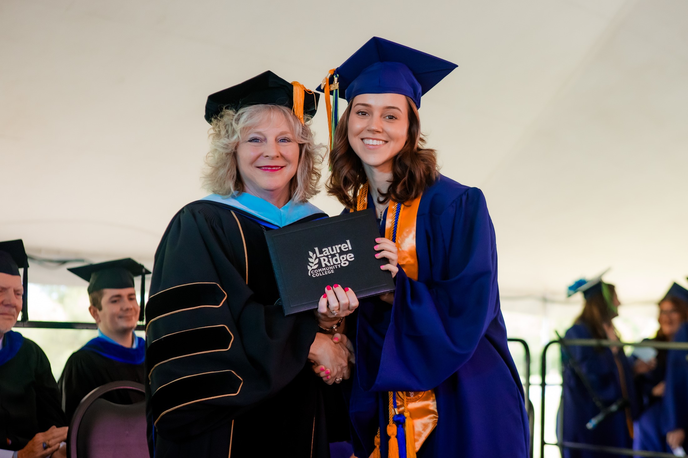 A graduate receives her diploma from Kim Blosser during a Laurel Ridge Community College commencement ceremony, both smiling and shaking hands while holding the diploma cover together.