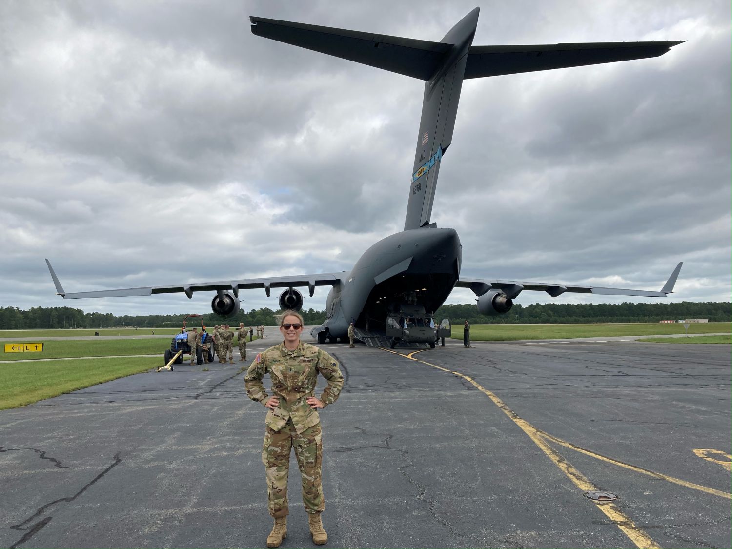 Valerie Harter stands on a tarmac in military camouflage uniform with hands on hips, smiling in front of a large military cargo aircraft with its rear loading ramp open and personnel working nearby.