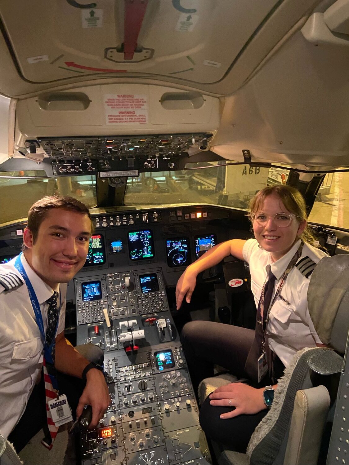 Valerie Harter and a fellow pilot smile from the cockpit of a commercial aircraft, both wearing pilot uniforms with epaulettes and lanyards while seated at the instrument-filled flight deck.